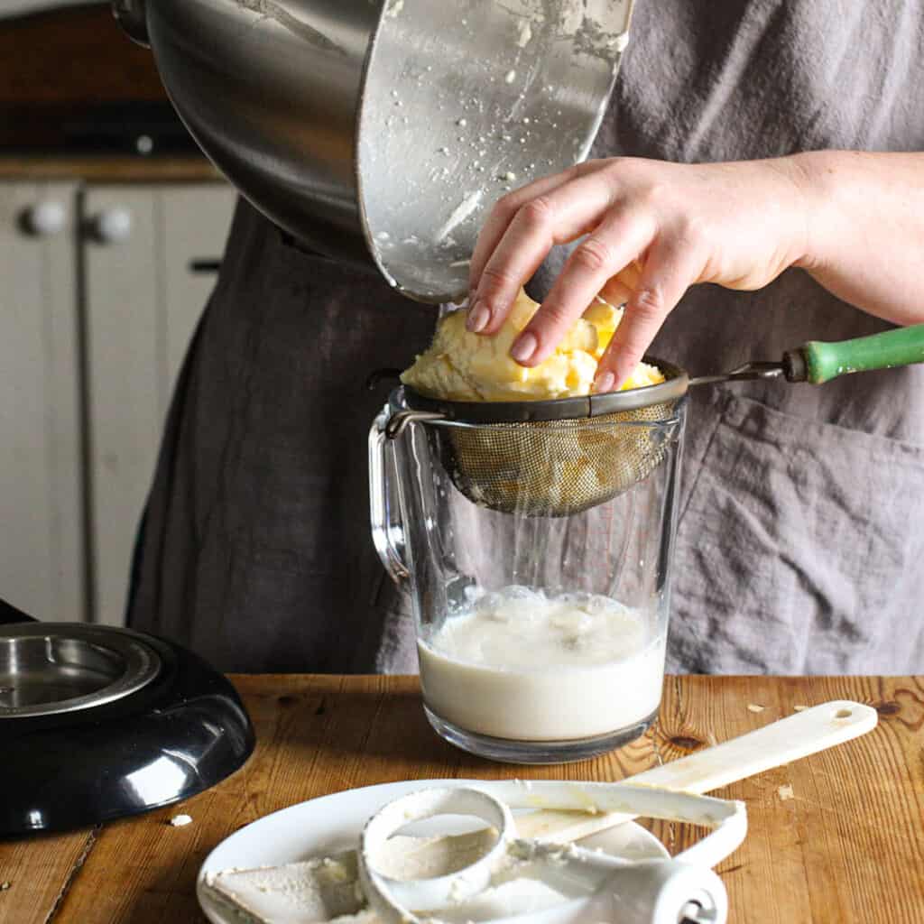 Woman in grey placing a large lump of fresh butter into a sieve allowing the buttermilk too drain into a the glass jar below 