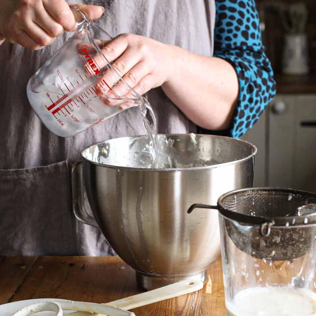 Woman pouring iced water into a large stainless steel mixing bowl from a glass jug