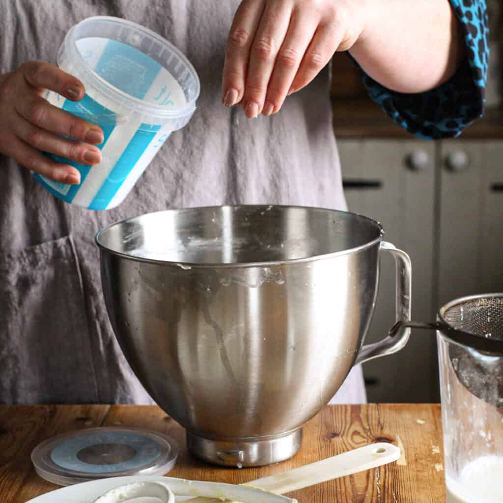 Woman in grey sprinkling salt form a large plastic tub into a stainless steel mixing bowl