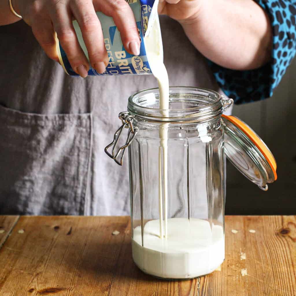 Woamns pouring double cream from a plastic tub into a large glass jar to make butter