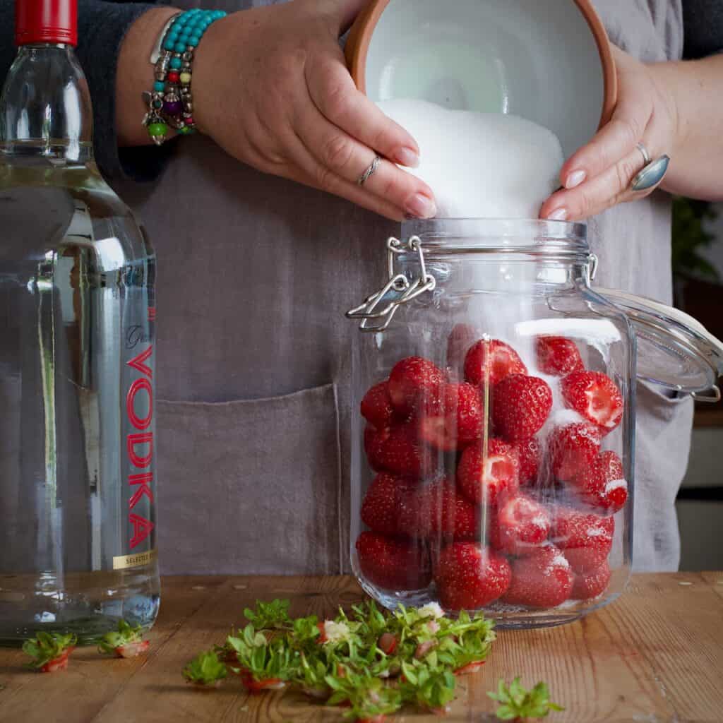 Woman tipping white sugar from a bowl into a glass jar of fresh strawberries