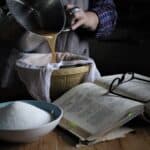 Woman pouring rosehip juice from a large silver saucepan into a bowl lined with a sieve with an old cookbook and bowl of sugar