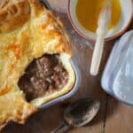 Venison meat pie peeping through a pastry shell with a bowl of beaten eggs with a pastry brush