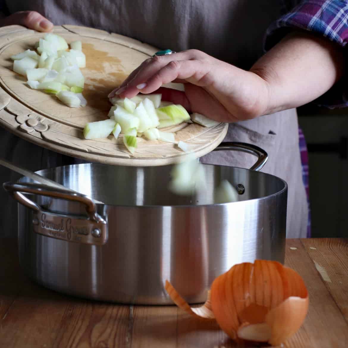 Womans hands scraping chopped onions from a wooden chopping board into a silver saucepan
