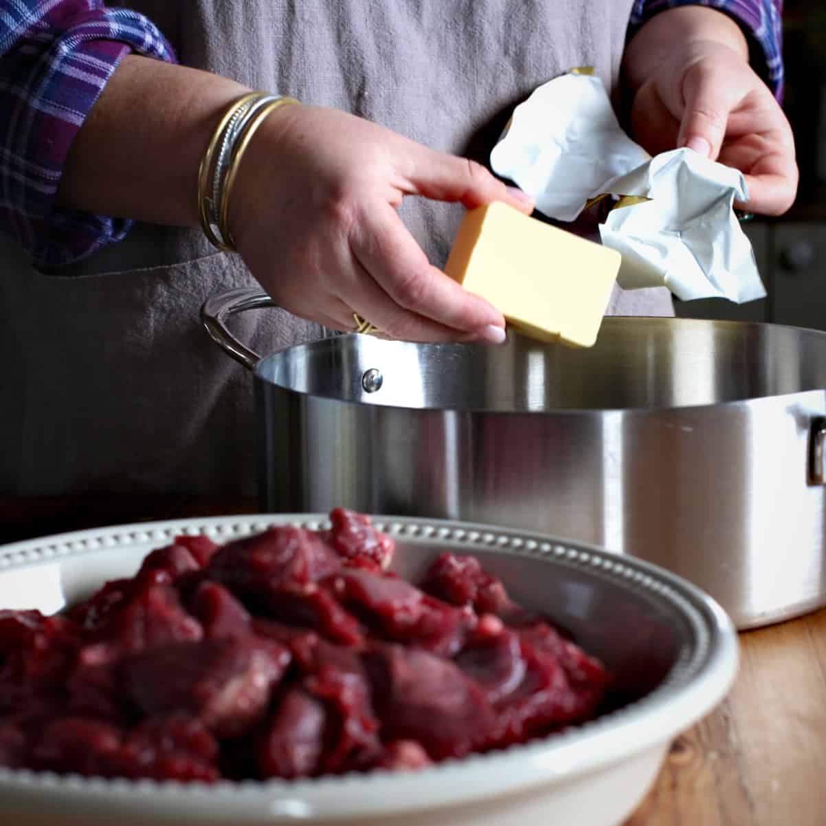 Woman placing a large lump of butter into a silver saucepan behind a white bowl of diced venison meat