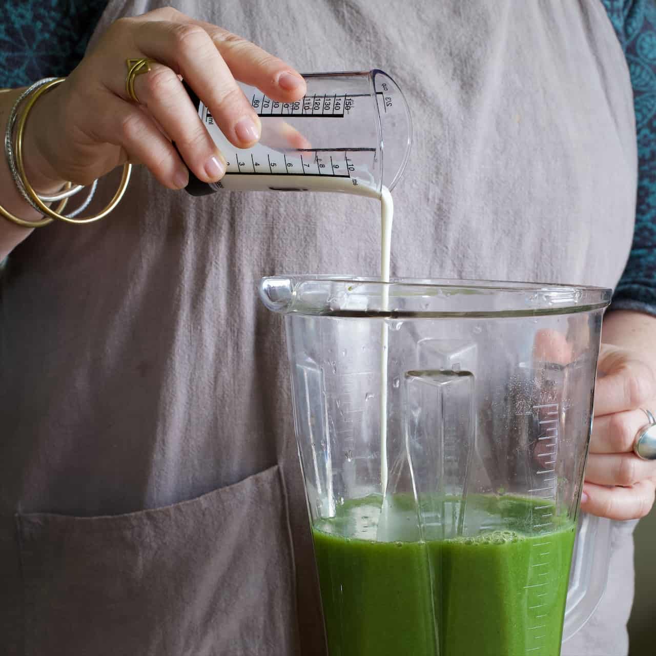 Woman in grey pouring cream from a clear plastic measuring jug into a blender jug of bright green nettle soup