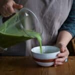 Womans hands pouring hot cream of nettle soup from a blender jug into a white serving bowl