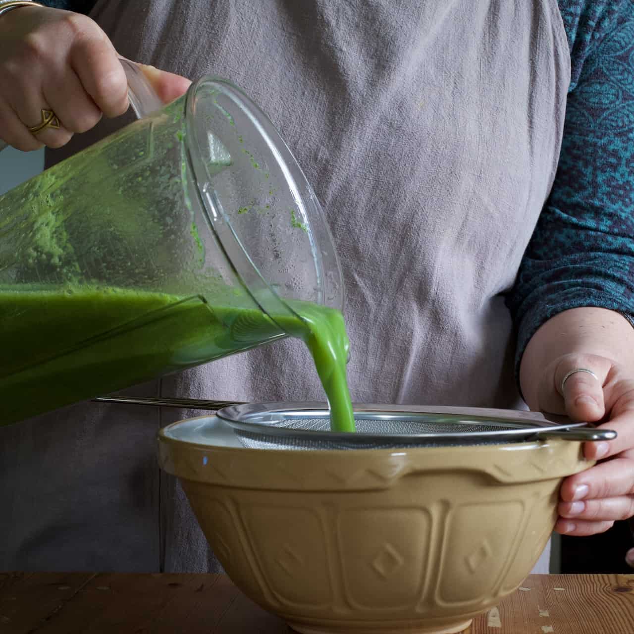 Woman pouring bright green soup from a clear plastic blender jug into a silver sieve balanced over a stone covered mixing bowl