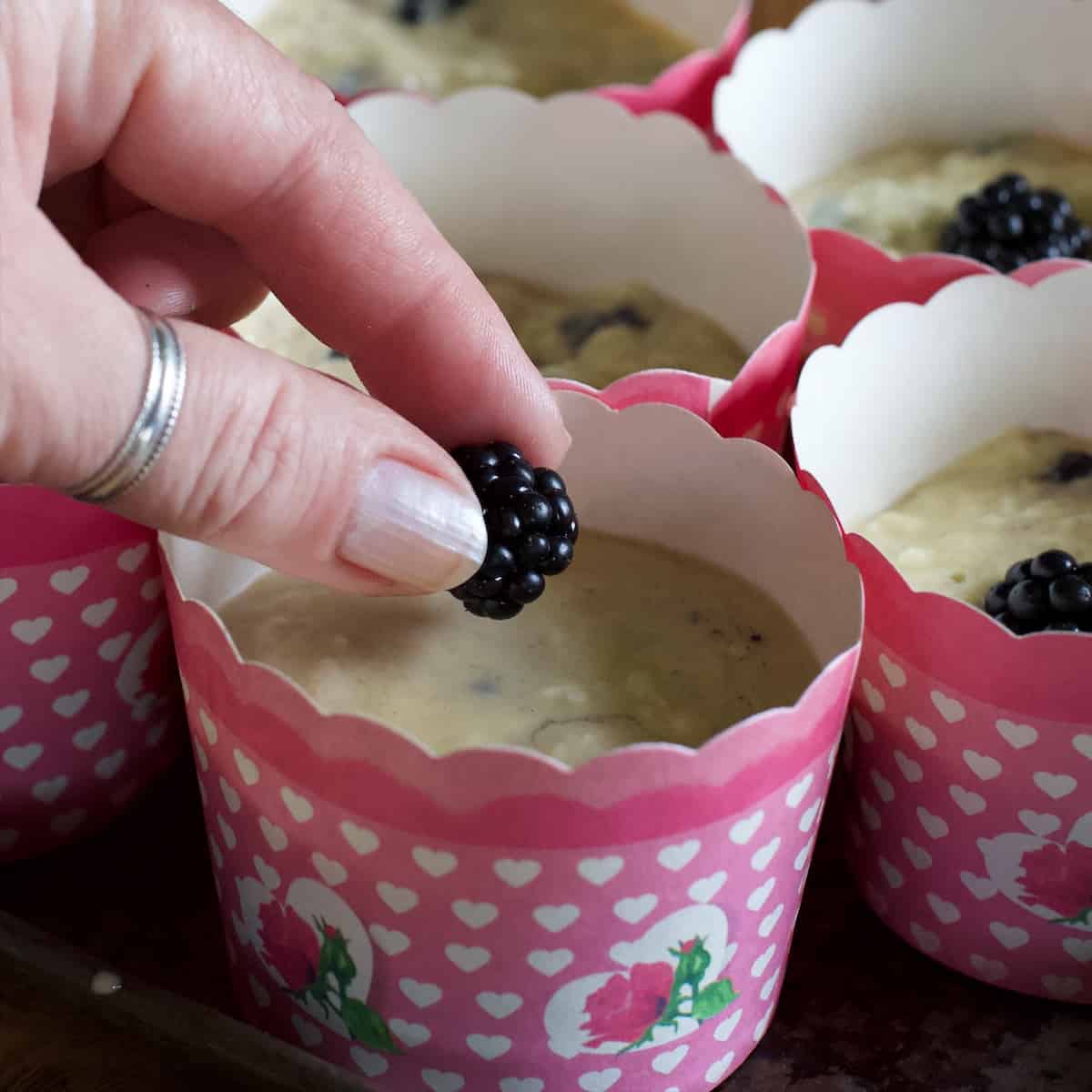 Woman placing a fresh blackberry on top of a muffin case filled with blackberry muffin batter