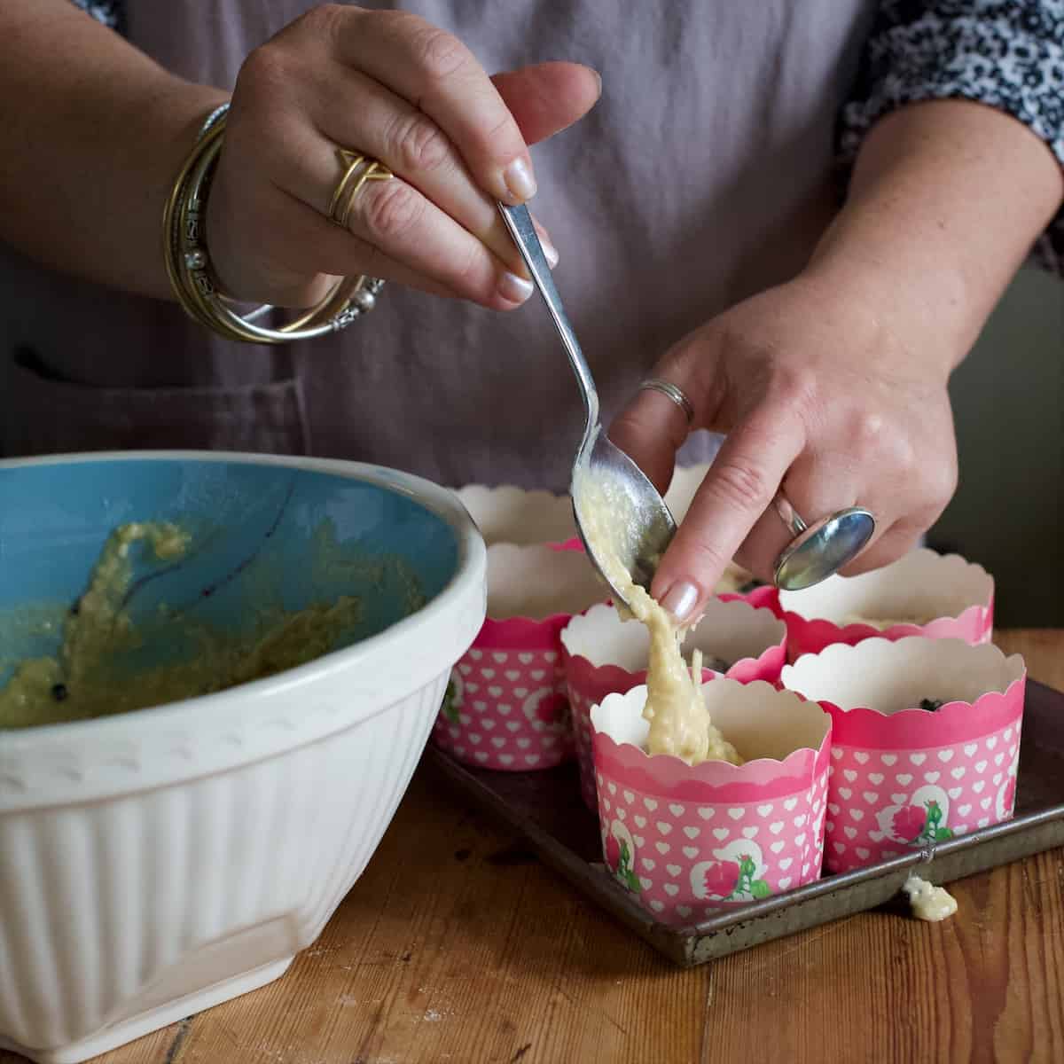 Woman spooning muffin batter from a blue and white mixing bowl into individual pink cupcake cases