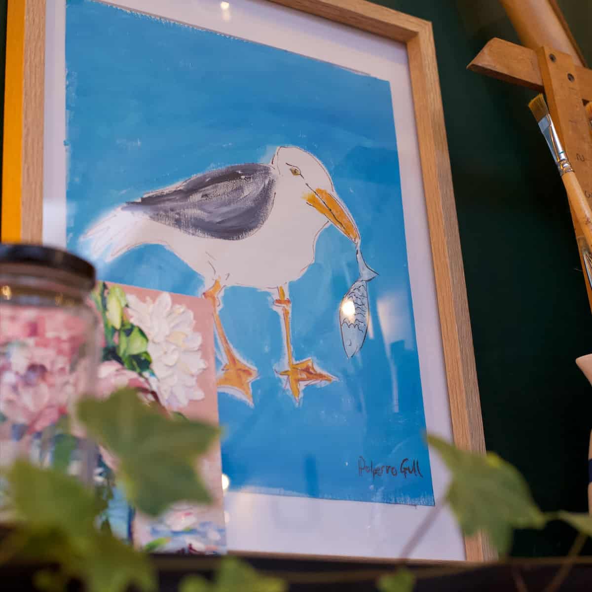 Painting of a seagull eating a fish in a bright naive style on a shelf with green ivy leaves