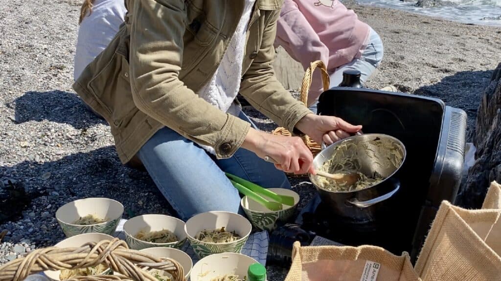 Woman on the beach serving noodles with fresh wild plants from a silver pan into 6 white bowls