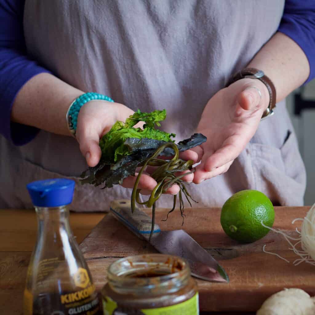 Woman in a rustic kitchen surrounded by fresh bright ingredients holding a range of freshly harvested seaweeds in her hands ready to cook with them