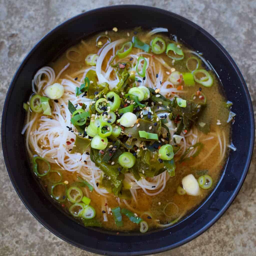 Close up shot of black bowl filled with rice noodles, miso broth, spring onions and seaweed