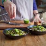 Womans hands sprinkling toasted sesame seeds onto a black bowl filled with a colourful mix of white noodles and green cooked seaweed on a wooden kitchen counter