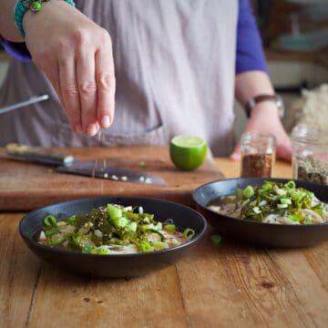 Womans hands sprinkling toasted sesame seeds onto a black bowl filled with a colourful mix of white noodles and green cooked seaweed on a wooden kitchen counter