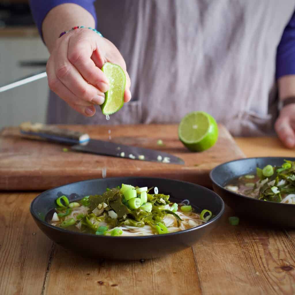Woman in grey squeezing a fresh lime over a black bowl of noodle and seaweed broth in a rustic kitchen setting