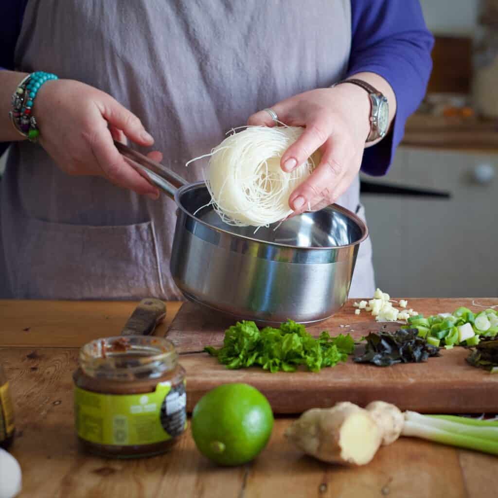 Woman in grey placing a block of dried rice noodles into a silver saucepan behind a a kitchen counter filled with fresh, vibrant ingredients such as lime, fresh seaweeds and miso paste.