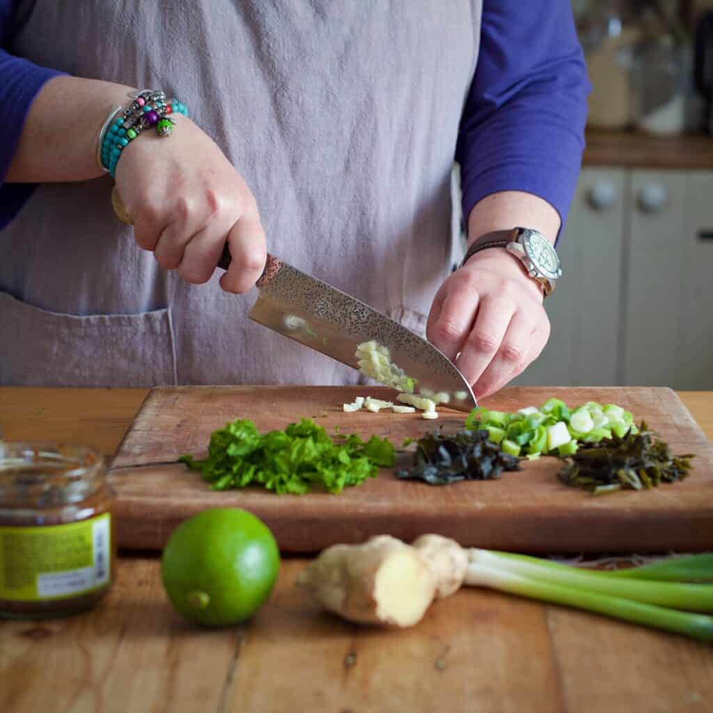 Woman in grey chopping garlic on a wooden board in a rustic kitchen surrounded by fresh ingredients