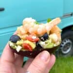 Womans hand holding a avocado shell stuffed with fresh basil, prawns, black olives and feta cheese